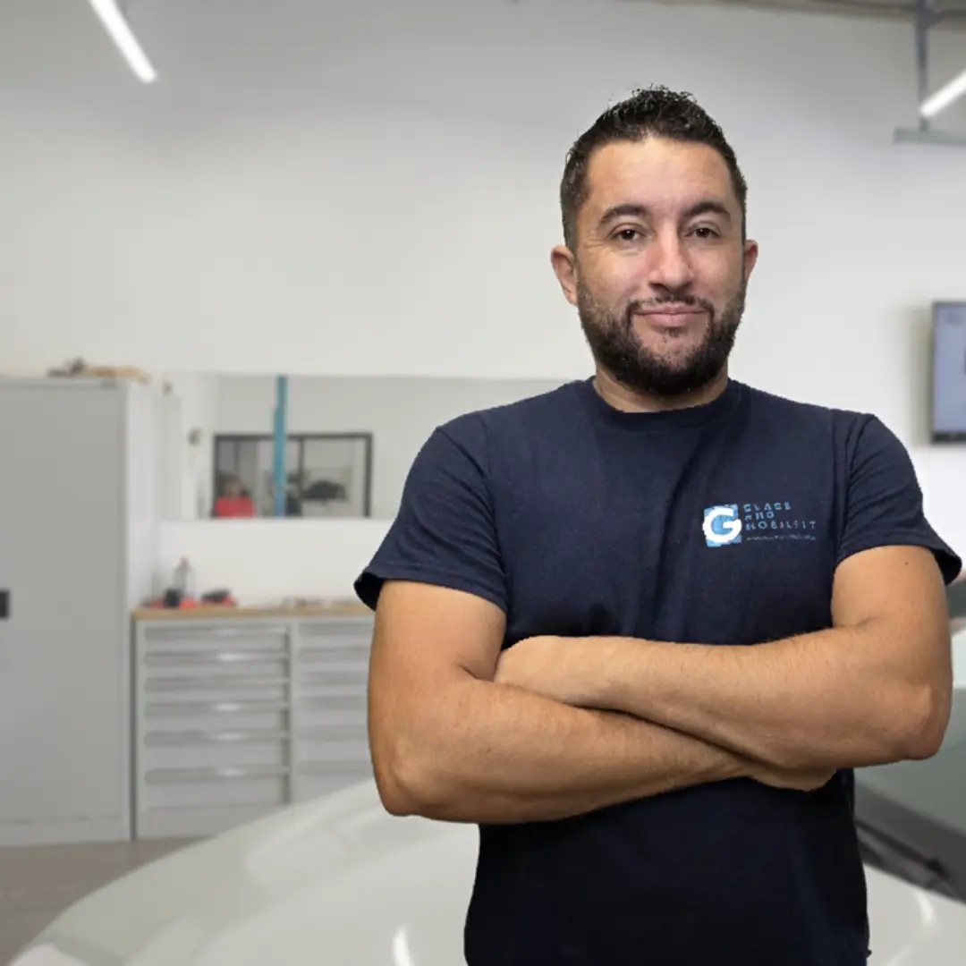 Yassine souriant, les bras croisés, portant un t-shirt bleu avec un logo "Glass Mobility", posant devant l'avant d'une voiture blanche dans un garage de remplacement de pare-brise moderne et lumineux, avec un léger effet de flou en arrière-plan